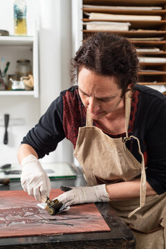 Mature female artist in apron and gloves covering engraved plate with ink while creating artwork in professional workroom