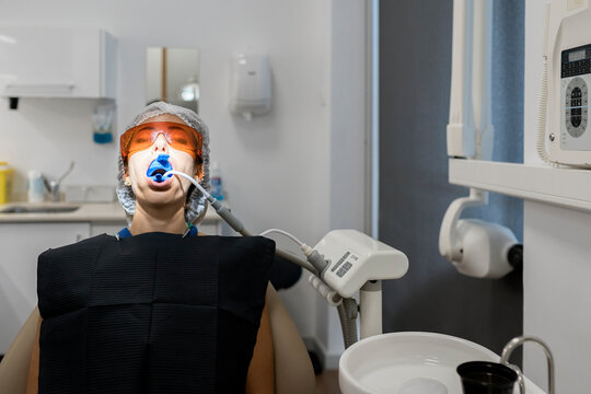 Female patient in laser safety goggles with dental expander and saliva ejector in mouth getting treatment procedure in modern clinic