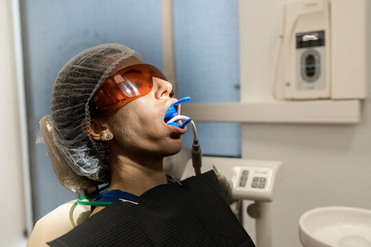 Female patient in laser safety goggles with dental expander and saliva ejector in mouth getting treatment procedure in modern clinic