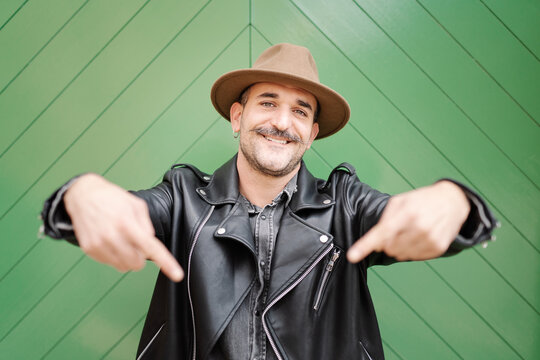 Trendy friendly male in hat and leather jacket smiling and looking at camera on green gate background