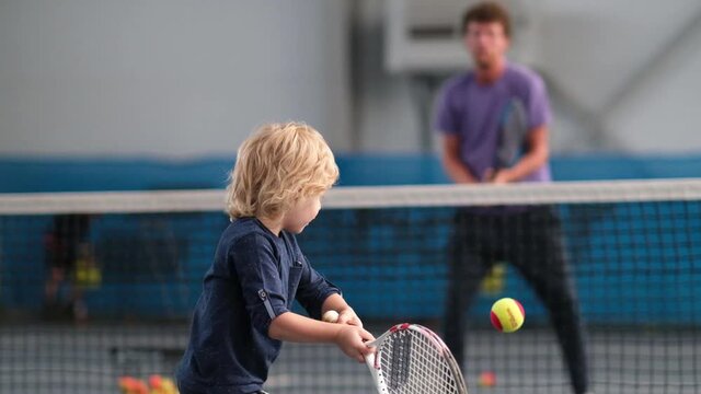 A man coach teaches a little boy to play on an indoor court. A professional tennis instructor throws the ball to hit the racket to the kid, how to return the ball with the racket
