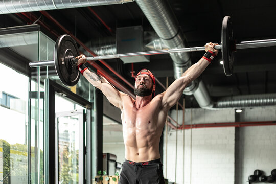 Muscular focused shirtless male athlete doing clean and jerk exercise with barbell during weightlifting training in gym looking away