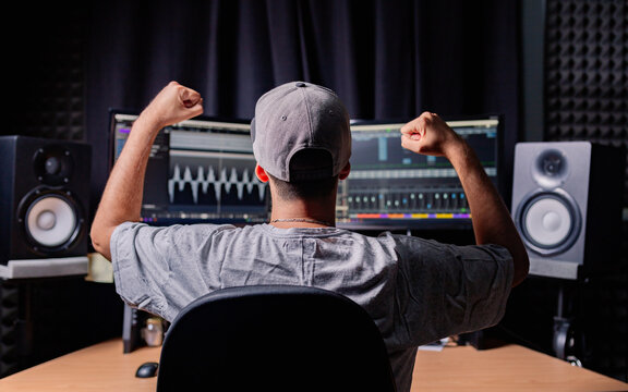 Back view of unrecognizable male musician sitting at table with monitors and stereo speakers while celebrating victory with fists up and recording audio in studio