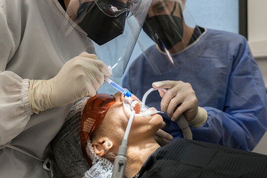 Crop unrecognizable dentist in protective mask and face shield making injection to patient while preparing for dental treatment in clinic with assistant during coronavirus pandemic