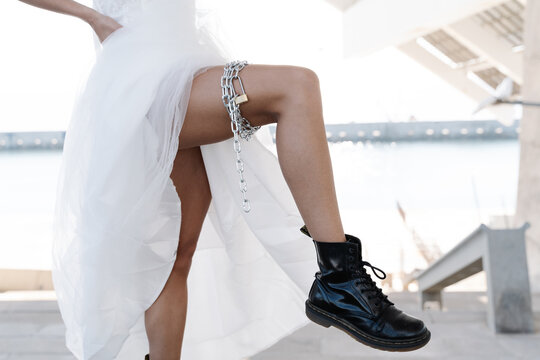 Crop unrecognizable female in elegant white wedding dress lifting leg in boots showing chain garter