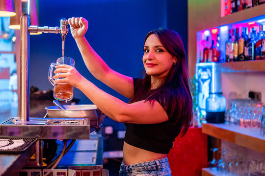 Side view of happy young female barkeeper pouring beer into glass while working in bar looking at camera