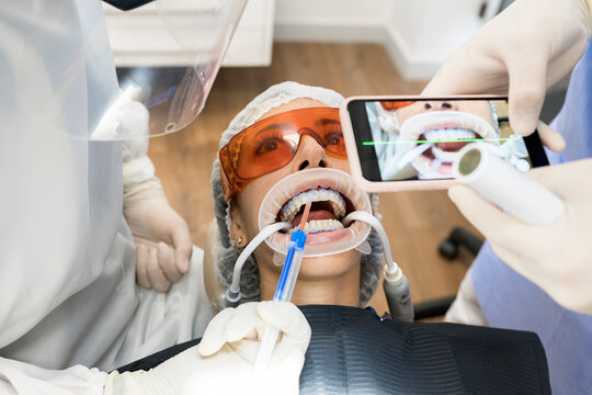 From above crop unrecognizable dentist taking picture of teeth of patient on smartphone while giving injection to patient preparing for dental treatment in clinic with assistant during coronavirus pandemic