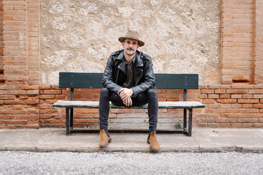 Serious male with mustache wearing hat and leather jacket relaxing on bench and looking at camera near stone brick wall