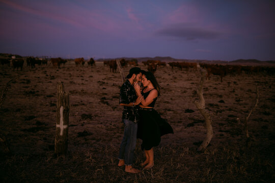 Full body side view of dreamy barefoot loving couple in casual clothing hugging with eyes closed r while standing in meadow under vibrant blue pink sky at sunset