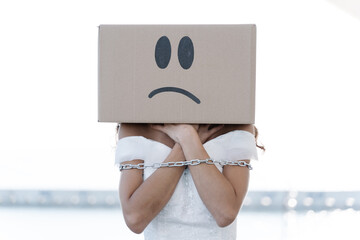 Anonymous female in white bridal dress with metal chain tying arms wearing cardboard box with unhappy smiley