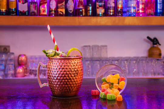Moscow mule cocktail with straw served with lime and mint leaves on counter near glass of marmalade sweets