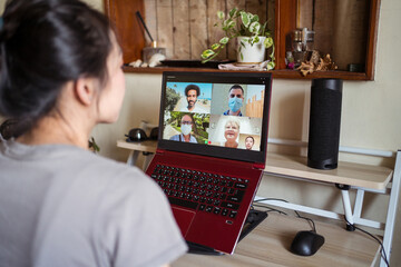 Young Asian female sitting on desk chatting via video call on laptop with group of friends while spending time at home during coronavirus pandemic