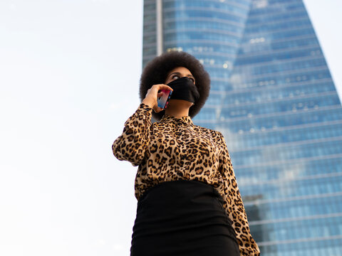 Low angle of confident African American female in protective mask having talk on mobile phone on city street