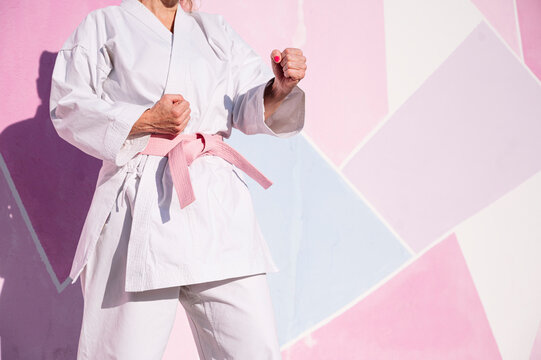 Cropped unrecognizable determined mature woman in pink head cover and belt fighting karate in cancer battle concept in the street on pink wall