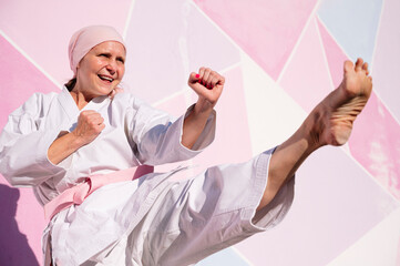 Determined cheerful mature barefooted woman in pink head cover and belt kicking fighting karate in cancer battle concept in the street on pink wall looking away