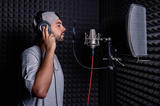 Side view of male singer in headphones and hat standing in acoustic room with soundproof walls and microphone and recording song in studio