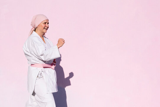 Side view of determined mature woman in pink head cover and belt fighting karate in cancer battle concept in the street on pink wall looking away