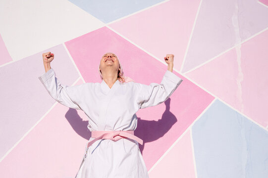Determined mature woman in pink head cover and belt fighting karate in cancer battle concept doing victory gesture in the street on pink wall looking up