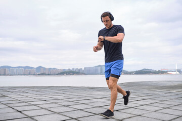 Serious male in headphones running along embankment and checking fitness tracker during cardio training