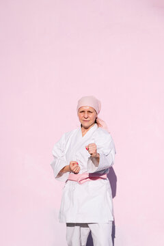 Determined mature woman in pink head cover and belt fighting karate in cancer battle concept in the street on pink wall looking at camera