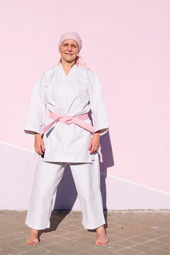 Determined mature woman in pink head cover and belt fighting karate in cancer battle concept in the street on pink wall looking at camera