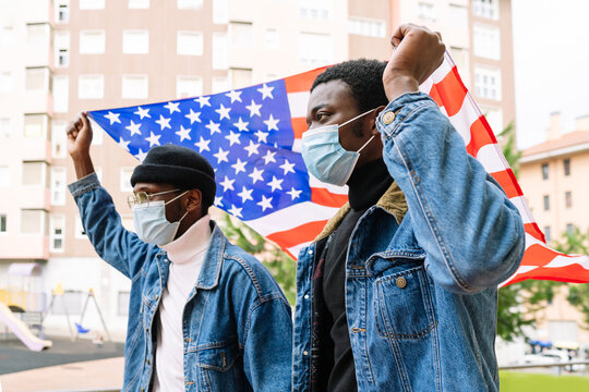 Low angle of cheerful African American male friends in masks standing with USA national flag on street and expressing joy