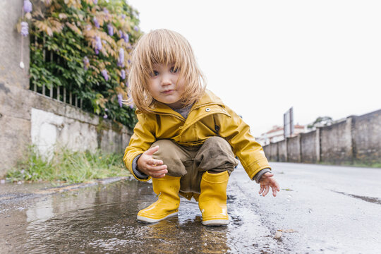 Low angle of adorable little child in yellow raincoat playing in puddle and splashing water while entertaining after rain on street looking at camera