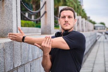 Concentrated male athlete in sportswear and headphones standing on street and warming up arms before workout in city