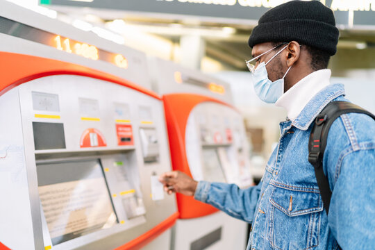 Side view of African American male traveler in mask buying ticket in self service machine on railway station during coronavirus pandemic