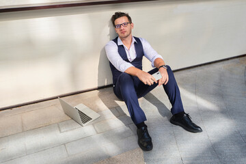 From above of young masculine office worker in formal suit and eyewear sitting on floor near wall with cellphone and netbook