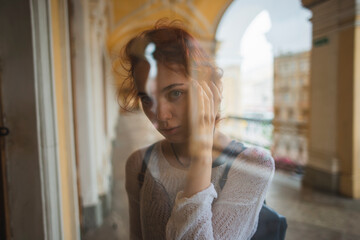 Through glass of young female in casual clothes with backpack standing on passage near historic building and looking at window in Saint Petersburg in Russia