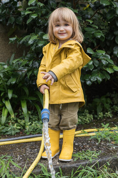 Cute little child wearing yellow raincoat and rubber boots watering garden from hose after rain and looking at camera