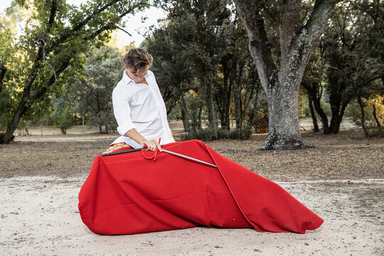 Serious slim male bullfighter in red pants and white shirt holding cloak performing gracefully before corrida in nature