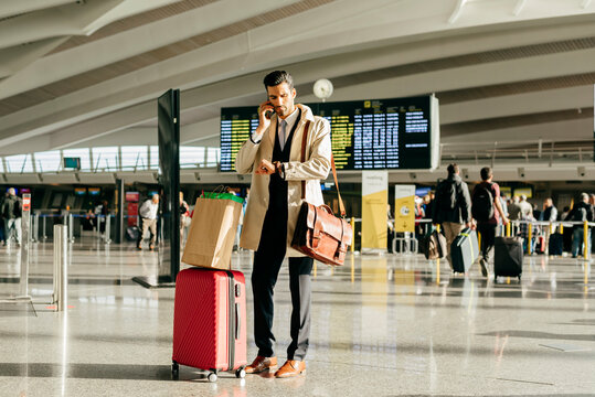 Man with dark hair in stylish clothes talking on phone and looking at watch while standing with suitcase in terminal of airport - Powered by Adobe