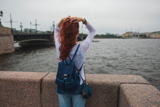 Back view of unrecognizable red haired female tourist with backpack standing near stone fence against river and enjoying wind while resting during sightseeing in cloudy day in Saint Petersburg in Russia