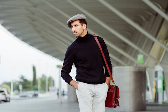 Fron view of man with beard in trendy clothes walking on street near asphalt road with parked cars at daytime