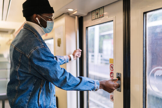 Side view of African American male passenger in mask standing at doors and pressing button before leaving train while traveling during coronavirus epidemic