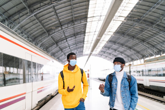 African American traveling males wearing protective masks walking along railway platform and talking before departure