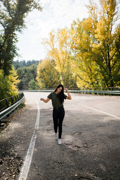 Full body of optimistic young female in casual outfit having fun and dancing on empty asphalt roadway while spending sunny autumn day in countryside