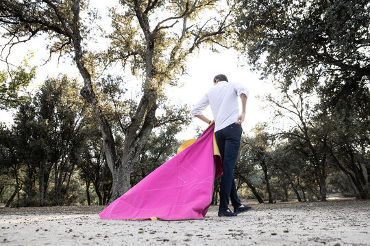 From below back view of unrecognizable graceful male matador practicing on meadow with yellow and pink cloak on sunny day