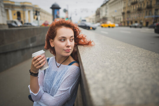 Positive millennial redhead female with cup of takeaway coffee leaning back on stone parapet and looking away thoughtfully while resting on street of Saint Petersburg in Russia