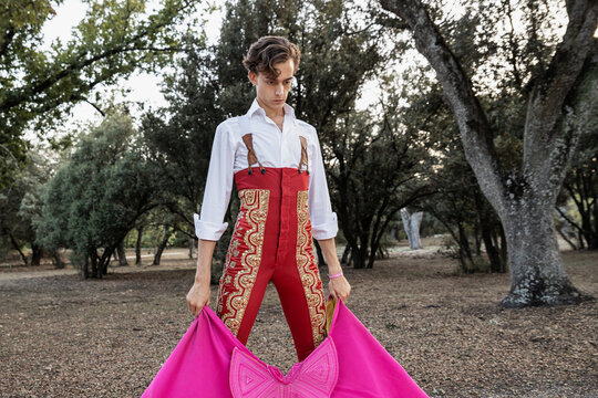 Slim focused thoughtful male bullfighter in red pants and white shirt holding cloak performing gracefully before corrida in nature