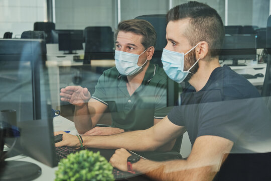 Male coworkers in medical masks sitting together using program for software during coronavirus pandemic