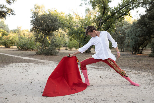 Side view of slim male bullfighter in red pants and white shirt holding cloak performing gracefully before corrida in nature
