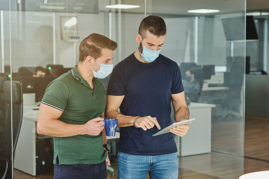 Busy male programmers in medical masks standing with tablet in office and discussing software development at work during coronavirus outbreak