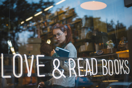 Through window side view of young stylish female in eyeglasses sitting in cozy bookshop with volume and looking at camera
