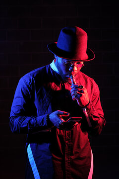 Confident African American male in stylish outfit and sunglasses smoking vape in studio with neon lights while using smartphone