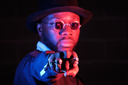 Serious African American male in stylish black clothes showing fist with rings at camera while standing on black background in studio with neon illumination