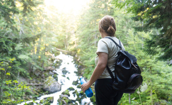 Back view of unrecognizable woman traveler with backpack standing on slope with bottle drinking water while admiring amazing landscape of green forest with fast river