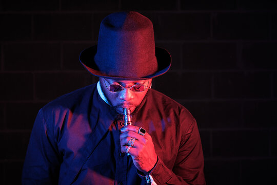 Stylish African American male in hat and sunglasses standing on black background in studio with neon illumination and smoking vape